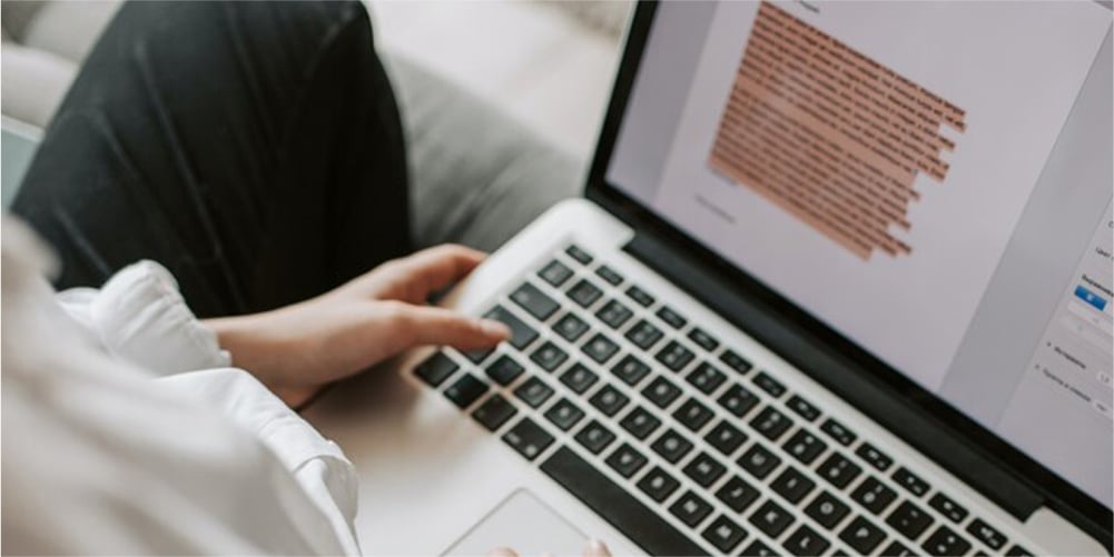 A woman is using a laptop on the bed, and the screen of her laptop displays highlighted text in a document file