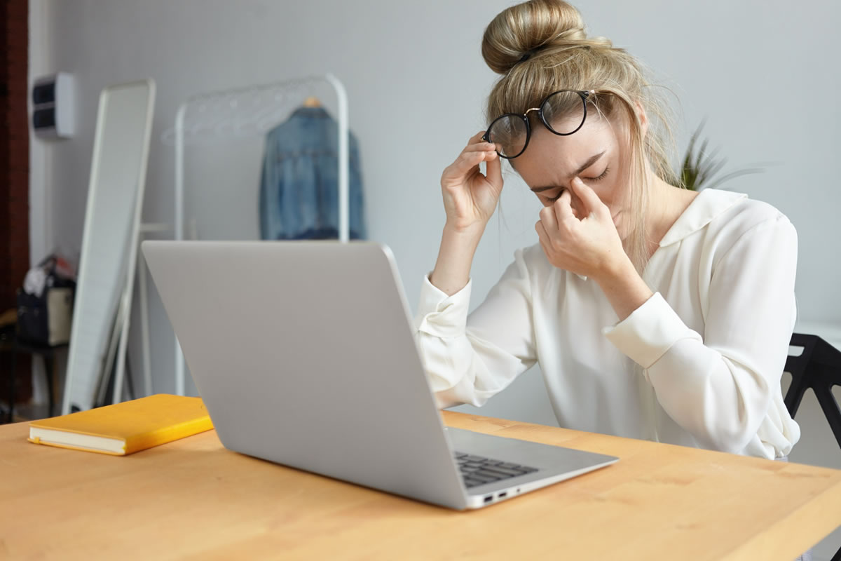 A girl removing her eyeglasses and pinching the bridge of her nose and a laptop infront of her