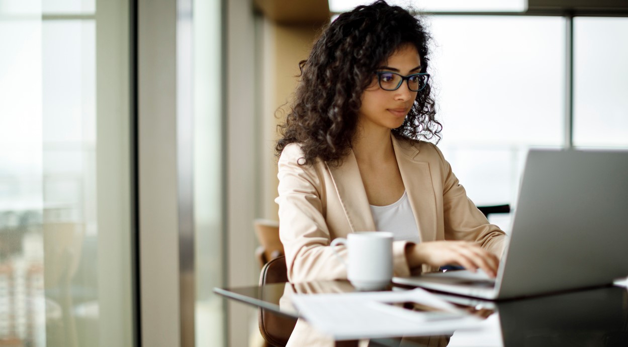 Woman with eyeglasses typing in a laptop with mug and papers beside her