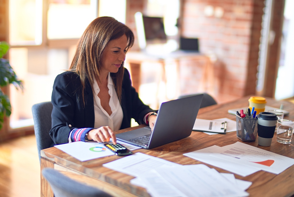 A girl in a suit typing in a laptop and using a calculator with papers and pens on the table