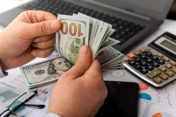 A manager of accounts counts cash in front of a laptop and a calculator