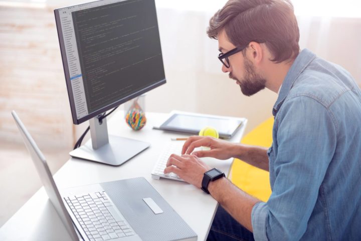A man concentrates on his monitor while a laptop is beside him on the table
