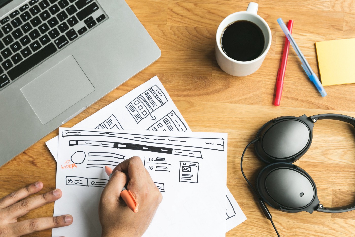 A man's hand writing a confusing content hierarchy on a piece of paper in front of his laptop, with a coffee cup, ballpoint pens, notes, and headphones nearby