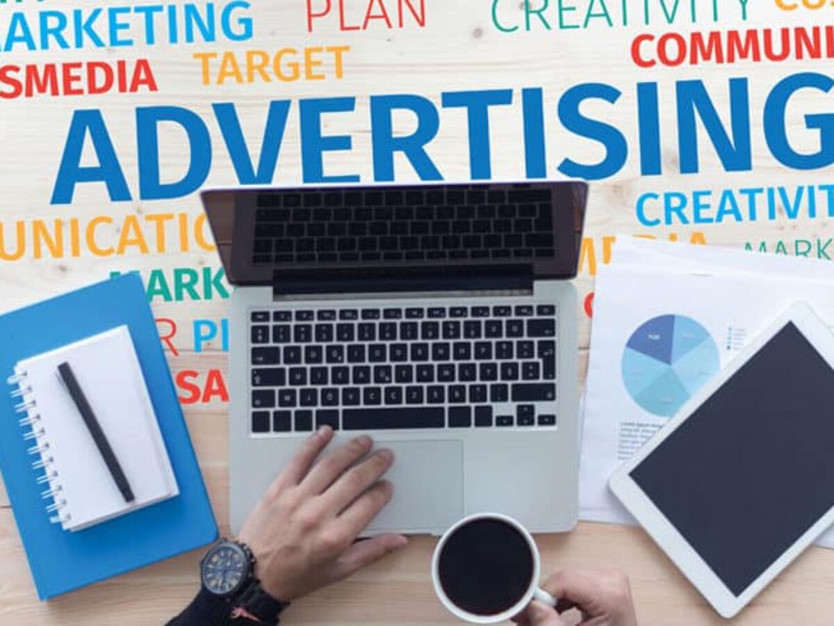 Hand in a laptop with notebook and tablet beside it and the word advertising behind it
