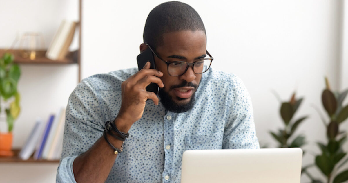 A man in blue polo talking on the phone while looking at a laptop