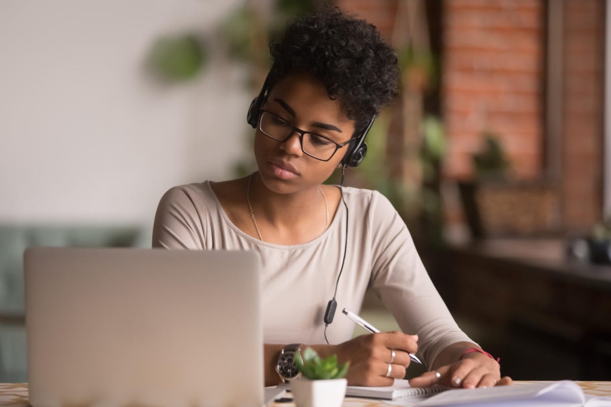 A woman in light brown long sleeves wearing a headset while holding a pen with a laptop on the table