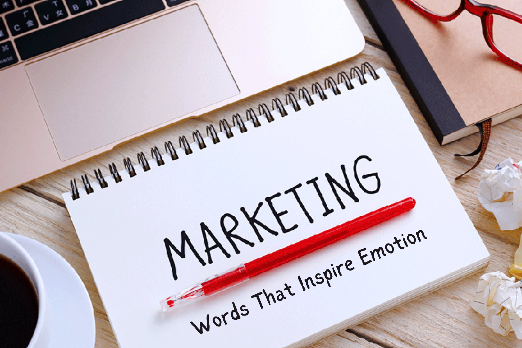 A notebook with the words "Marketing Words that inspire emotion" written on top of a table with a laptop and a cup of coffee