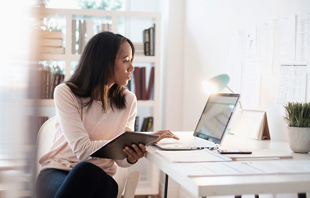 A woman in white long sleeves holding a tablet while looking at the laptop in front of her