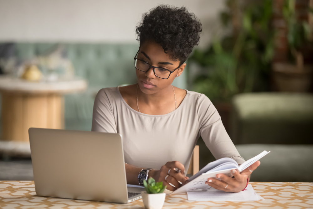 A woman holding a notebook while looking at the laptop in front of her