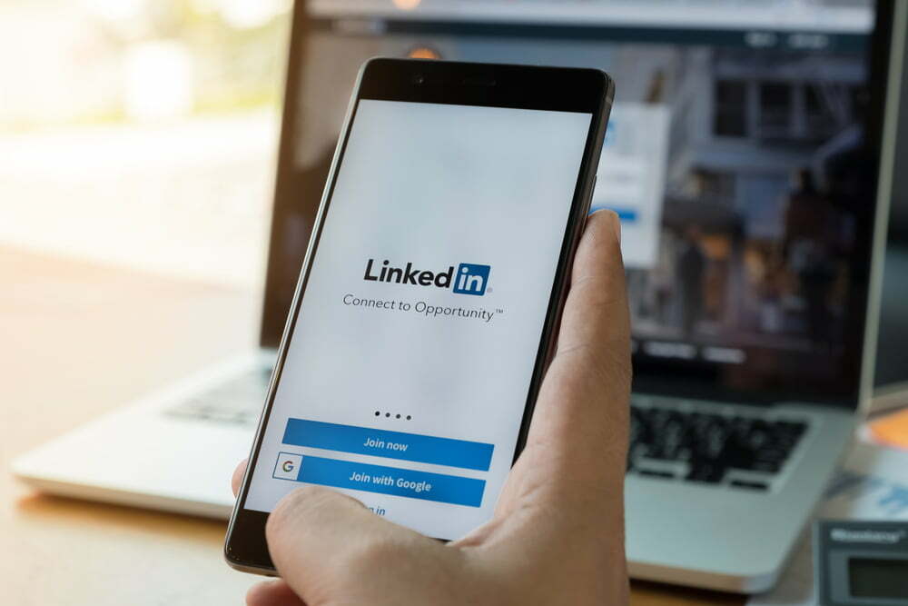 Hand holding a phone with LinkedIn application and laptop behind it